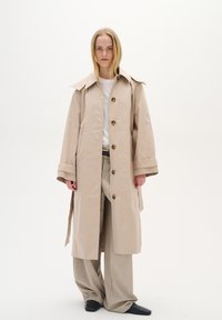 Beige trenčkot s širokým límcem, předním zapínáním na knoflíky a dlouhými rukávy. Kombinovaný se světle zbarvenými kalhotami a černými botami.