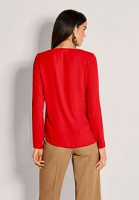 Frau mit langen dunklen Haaren, die ein rotes Langarmoberteil und beige Hosen trägt, steht mit dem Rücken zu einer einfachen hellen Hintergrund.