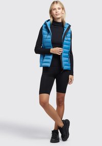 Kvinna står med ett ben korsat, iklädd en blå ärmlös pufferväst över en svart polotröja, svarta shorts och svarta sneakers.