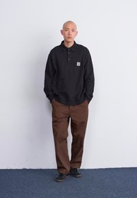 Polo a maniche lunghe nera con colletto e logo a toppa, abbinata a pantaloni larghi marroni e scarpe nere, su sfondo bianco.