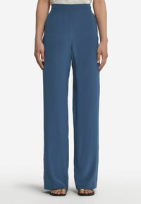 Blå vida byxor i ett mjukt material, med hög midja och full längd. Sömmen slutar vid vristen och de är kombinerade med enkla svarta sandaler.