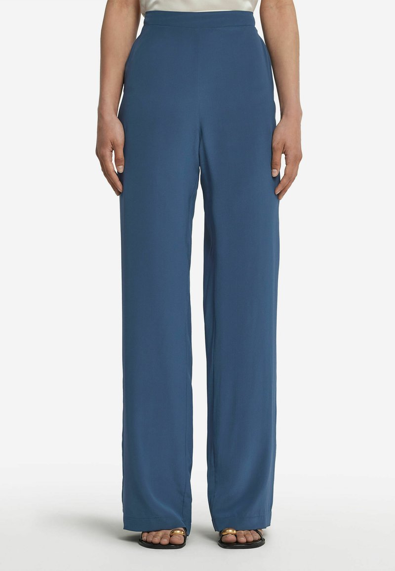 Blå vida byxor i ett mjukt material, med hög midja och full längd. Sömmen slutar vid vristen och de är kombinerade med enkla svarta sandaler.