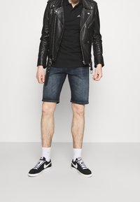 Černá kožená motorkářská bunda, černé polo tričko, tmavé džíny šortky a černé tenisky Nike s bílými akcenty, umístěné na jednolitém pozadí.