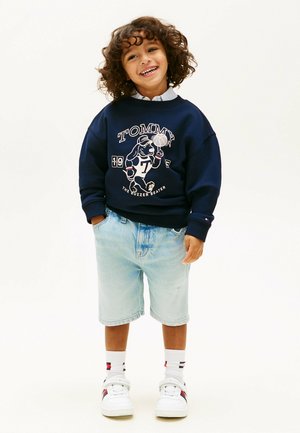 Uśmiechnięte dziecko z kręconymi włosami, ubrane w granatową bluzę z grafiką psa i koszykówki, jasne szorty dżinsowe, białe trampki i białe skarpetki, stojące.