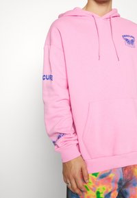 Sudadera rosa hecha de un tejido suave, con texto azul y gráficos en las mangas, y un bolsillo canguro en la parte delantera.