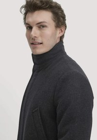 Dunkelgrauer Wollmischmantel mit hohem Kragen, Seitentasche, schlankem Design und strukturierter Oberfläche, getragen von einem Modell, das leicht nach links blickt.