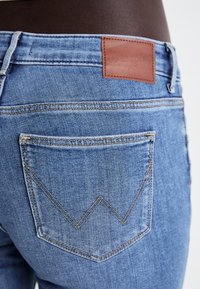 Jeans de denim azul claro com um rótulo de cinto em pele castanha. Apresenta um bolso traseiro com detalhes de costura em zigue-zague.