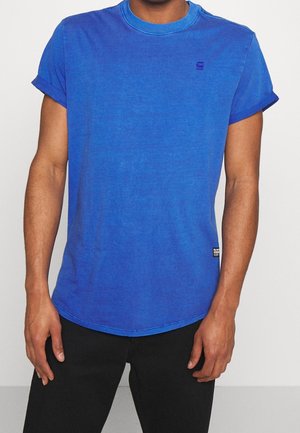 Mann trägt ein blaues T-Shirt mit kurzen Ärmeln und aufgekrempelten Säumen sowie schwarze Hosen, steht vor einem schlichten, hellen Hintergrund.