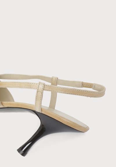 Sandale beige à bout ouvert avec des fines brides et un petit talon noir brillant sur fond blanc.