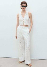 Witte cropped top met een kantpatroon en een strik aan de voorkant, gecombineerd met hoge, wijde broek met oogjesdetails. Zwarte zonnebril.