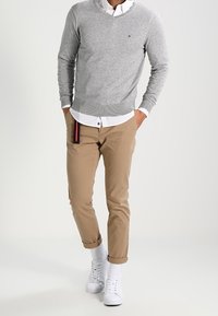 Suéter gris de cuello en V sobre una camisa blanca de cuello, combinado con chinos color marrón claro y zapatillas blancas. Logo notable en el suéter y detalle en el bolsillo.