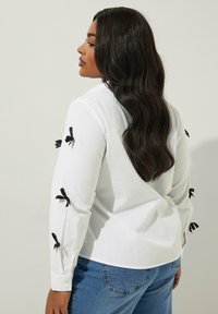 Camicia bianca a maniche lunghe con dettagli in nastro nero sulle maniche, caratterizzata da una texture liscia e una vestibilità morbida, abbinata a jeans blu.