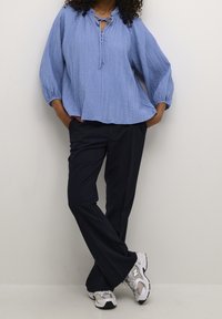 Blouse bleu clair texturée avec un nœud devant, coupe ample, associée à un pantalon large bleu marine foncé et des chaussures de sport grises.