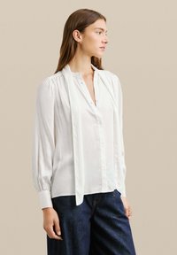 Blusa bianca con maniche lunghe e chiusura a bottoni, caratterizzata da un colletto morbido e un dettaglio di cravatta drappeggiata. Abbinata a pantaloni in denim scuri.