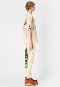 Camiseta oversize beige con estampados gráficos, combinada con pantalones cargo crema con detalles de bolsillos y una etiqueta negra, complementada con botas marrones de cordones.
