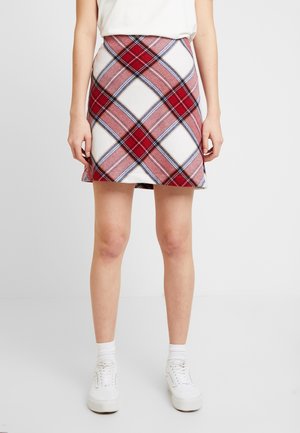 Kobieta ma na sobie czerwoną, różową i białą kratowaną mini spódnicę, białe skarpetki i białe sneakersy na platformie na jednolitym tle.