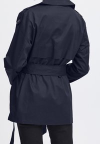 Tmí modrý trench coat s páskem, hladkým povrchem, širokým límcem a ramenními epauletami; má ozdobný detail na zadní části.