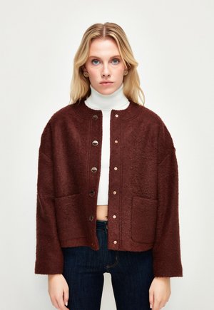 Braune, strukturierte Jacke mit Rundhalsausschnitt, Druckknöpfen und zwei vorderen Taschen; kombiniert mit einem weißen Rollkragenpullover und dunklen Jeans.