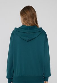 Tyrkysová oversized mikina s jemnou texturou, kapucí na šňůrku a žebrovanými manžetami. Design má volný střih a je vyroben z bavlněné směsi.