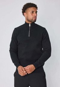 Pull noir à col zippé avec des poignets et un ourlet côtelés. Fabriqué en tissu tricoté, il présente une fermeture éclair argentée et un design ajusté.