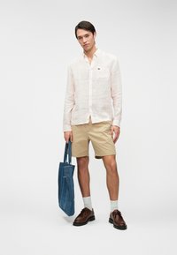 Chemise à manches longues rayée rose clair, shorts beiges, chaussures marron et sac fourre-tout en denim. Design simple et décontracté avec des coutures et textures visibles.