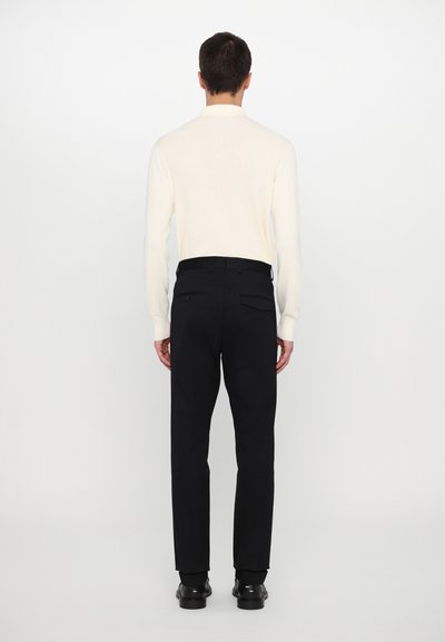 Polo de malha de manga longa em creme para homem, de design ajustado, combinado com calças pretas ajustadas, apresentando dois bolsos traseiros e calçado preto.