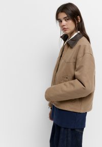 Brun bomuldsjakke med en brun læderkrave, med lynlås og sidelommer, stylet over en navy top og løse denimbukser.