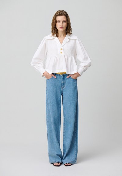 Blusa blanca corta con textura fruncida, frente con botones y mangas anchas, combinada con unos pantalones vaqueros de pierna ancha de cintura alta en azul claro y sandalias negras.