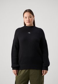 Pull en tricot côtelé noir avec un col montant et une coupe décontractée. Il présente un petit patch logo sur la poitrine et des manches larges.