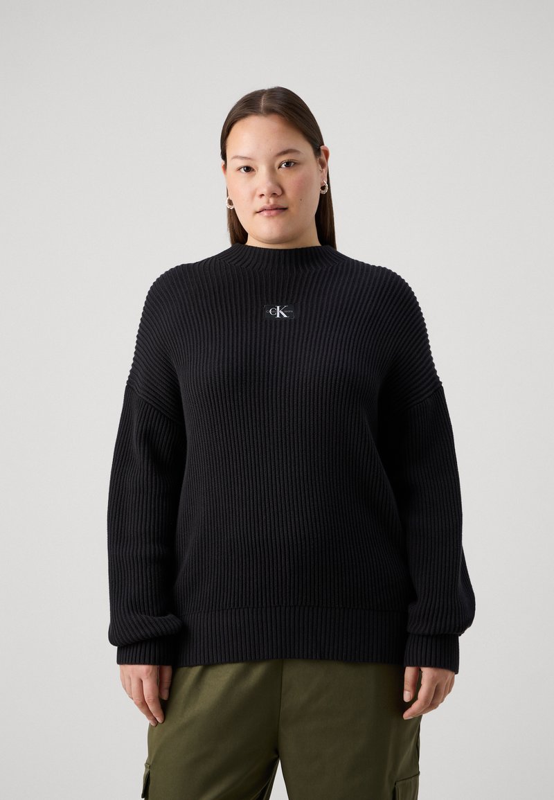 Pull en tricot côtelé noir avec un col montant et une coupe décontractée. Il présente un petit patch logo sur la poitrine et des manches larges.