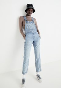 Šviesiai mėlynos džinsinės salopetės su reguliuojamomis petnešomis, dviem priekinėmis kišenėmis ir siaurėjančiomis kojomis, derinamos su rudomis ilgomis rankovėmis marškiniais ir juoda kibiro kepure.