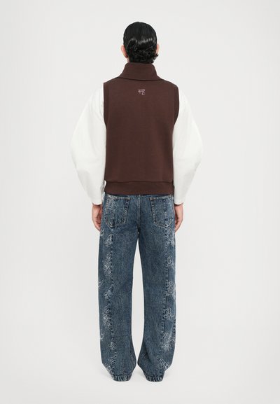 Brauner ärmelloser Pullover mit hohem Kragen, getragen über einem weißen Langarmhemd. Kombiniert mit blauen Denim-Jeans mit floralem Stickmuster.