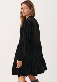 Abito in velluto nero con maniche lunghe, scollatura voluttuosa e orlo a strati. Tessuto morbido e forma fluente. Modella mostrata di lato.