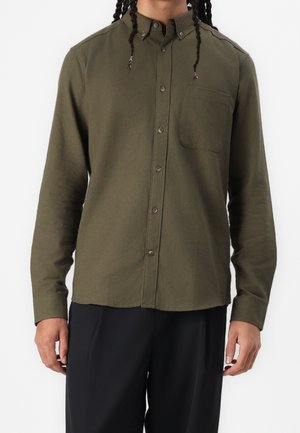 Homme portant une chemise boutonnée vert olive avec une poche poitrine et un pantalon noir, debout devant un fond uni.
