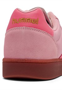 Sapatilha em camurça e pele rosa com um calcanhar texturizado rosa, com a marca "hummel" em dourado e uma sola de borracha.