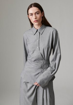 Femme aux longs cheveux bruns portant une chemise grise boutonnée et un pantalon ample assorti sur un fond gris uni.