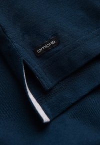 Námořnická modrá textilie se strukturovaným tkaním, která má černou etiketu s logem "ombre". Okraje mají kontrastní bílou úpravu.