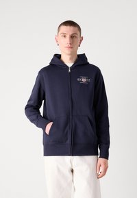 Marineblå zip-up hettegenser laget av bomullsblanding, med et logo foran, snøring i hetta, og to frontlommer. Glatt tekstur og avslappet passform.