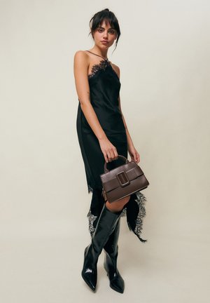 Kvinde iført en sort kjole med blonder og knæhøje sorte støvler, der holder en brun håndtaske mod en ensartet lys baggrund.