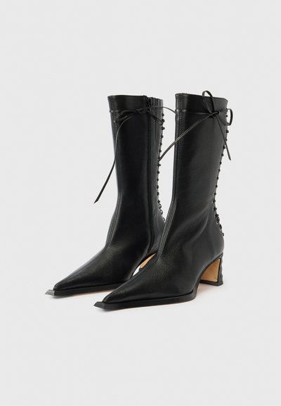 Μαύρες δερμάτινες μπότες μέχρι το γόνατο με μυτερή μύτη, κυρτή φτέρνα και διακοσμητικό κορδόνι στο πλάι. Λείο υλικό με ελάχιστες λεπτομέρειες.