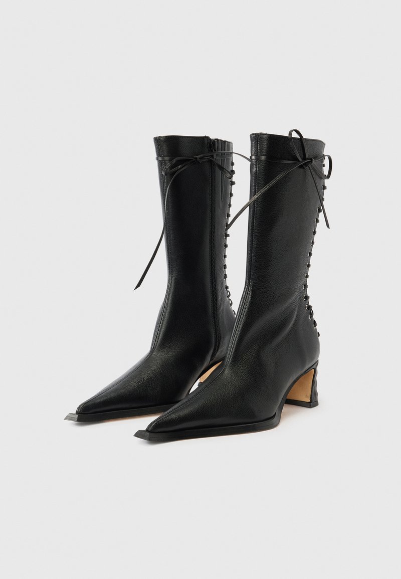 Schwarze kniehohe Stiefel aus Leder mit spitz zulaufender Schuhspitze, geschwungenem Absatz und seitlicher Schnürung. Glatte Textur mit minimalen Akzenten.