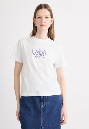 T-shirt en coton blanc avec un logo "Calvin Klein" violet sur le devant. Manches courtes, encolure ronde, coupe décontractée. Porté avec une jupe en denim bleue.
