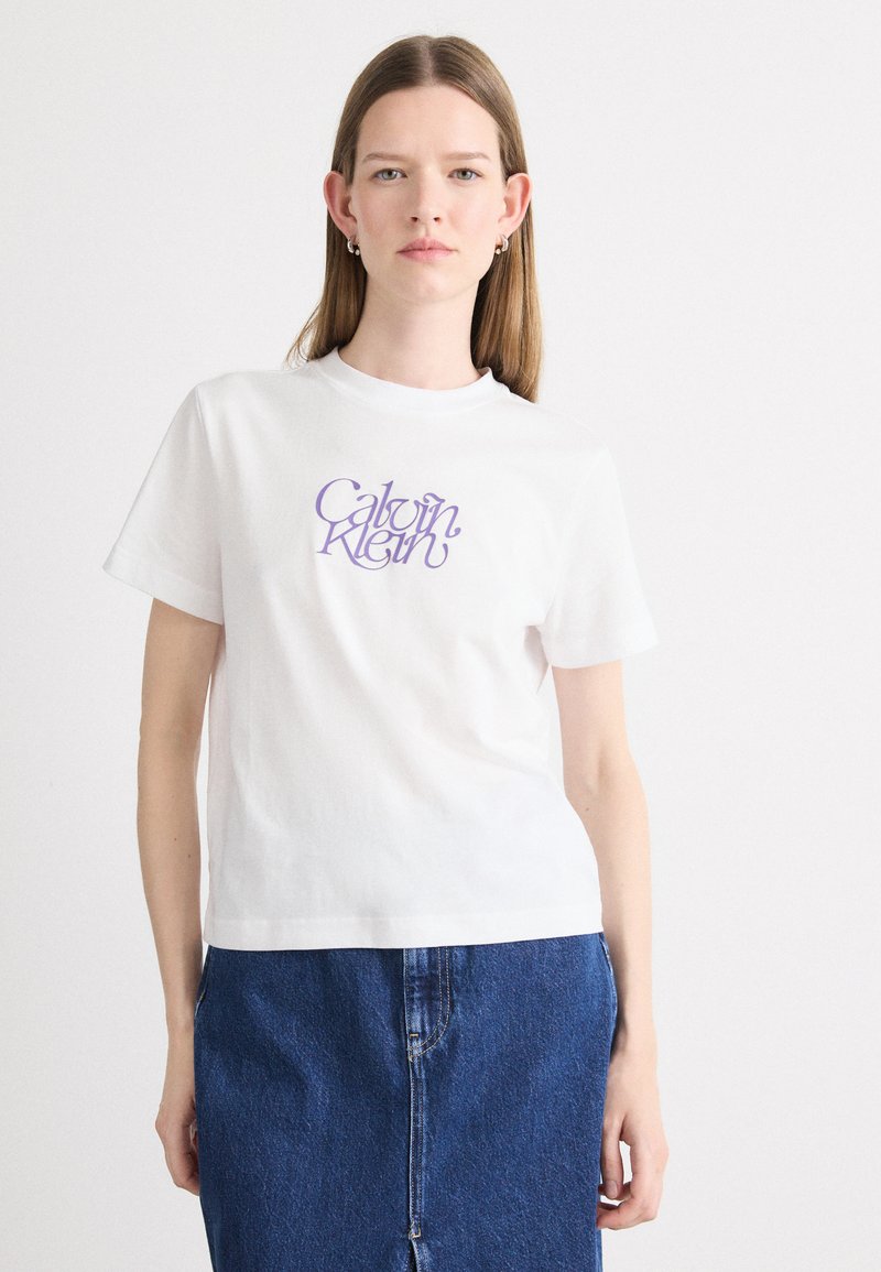 Witte katoenen t-shirt met paarse "Calvin Klein" logo op de voorkant. Korte mouwen, ronde halslijn, ontspannen pasvorm. Draagt met een blauwe denimrok.