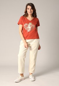 Roodachtig katoenen t-shirt met een goudkleurige hartafdruk van zebra's, gecombineerd met crèmekleurige ribfluwelen broek en witte sneakers.