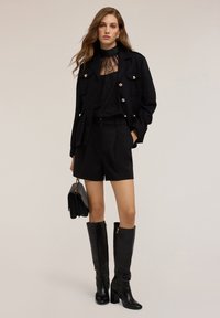 Giacca nera corta con bottoni dorati, top in pizzo, pantaloni shorts neri su misura, stivali neri testurizzati al ginocchio e una borsa nera.