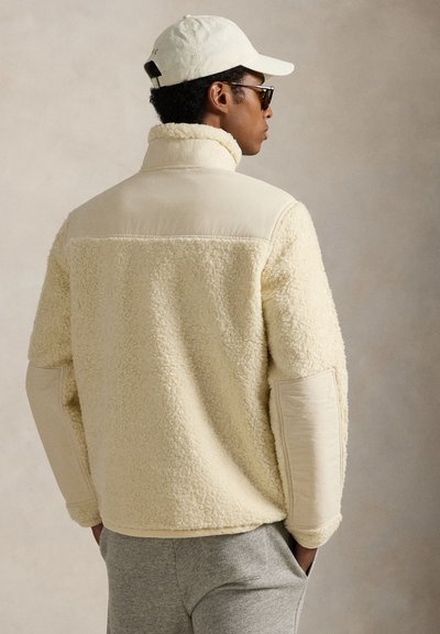 Kreemivärvi fliisjope tekstuurse, koheva kere, kontrastsete siledamate varrukate ja kõrge kraega. Ühendatud hallide pükste ja mütsiga.