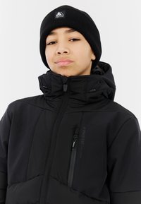 Schwarze isolierte Jacke mit hohem Kragen, Reißverschluss-Brusttasche und Kapuze. Getragen mit einer schwarzen gerippten Mütze mit einem Logo.