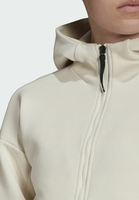 Krémová mikina s kapucí na zip, s jemnou texturou, zakulaceným límcem a kontrastním černým táhlem zipu.