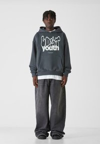 Felpa oversize grigio scuro con grafica bianca "LOST youth", tasca a marsupio, abbinata a jeans neri sbiaditi e larghi, e sneakers nere.