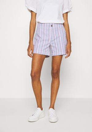 Kvinde iført hvide sneakers, multifarvede vertikale stribede shorts og en hvid top med korte ærmer mod en ensfarvet baggrund.